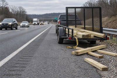 Lumber and a loose tie-down beside a utility trailer on a highway shoulder